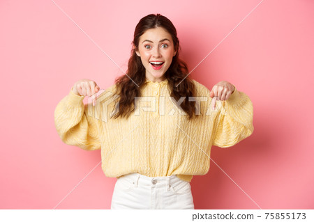 Excited cute girl showing advertisement, pointing fingers down and smiling at camera, demonstrating logo, standing against pink background 75855173