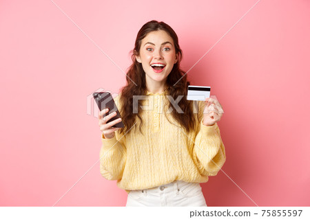 Technology and online shopping. Excited girl making order, paying online with plastic credit card, holding mobile phone and smiling at camera, pink background 75855597
