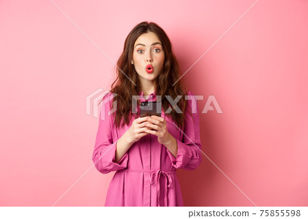 Image of surprised young woman gasping and saying wow, staring impressed at camera after reading news online on mobile phone, standing with smartphone against pink background 75855598