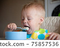 Focused adorable toddle in colorful bib sitting in highchair and eating porridge with spoon. 75857536
