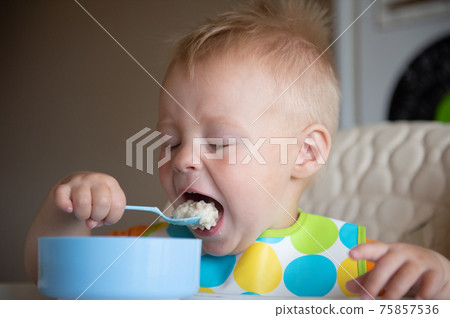 Focused adorable toddle in colorful bib sitting in highchair and eating porridge with spoon. Focused adorable toddle in colorful bib sitting in highchair and eating porridge with spoon. 75857536