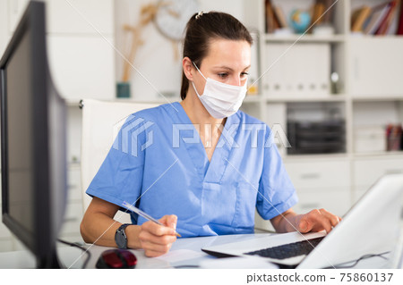 Woman doctor in protective mask sitting at workplace with computer in office Woman doctor in protective mask sitting at workplace with computer in office 75860137
