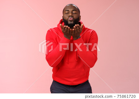 Charming cute african american bearded guy in red hoodie close eyes folding lips hold palms near mouth sending passionate air kiss blowing mwah camera, standing lovely pink background Charming cute african american bearded guy in red hoodie close eyes folding lips hold palms near mouth sending passionate air kiss blowing mwah camera, standing lovely pink background 75861720