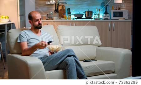 Bored man switching TV channels, holding popcorn in hads Bored man switching TV channels, holding popcorn in hads 75862289
