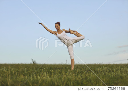 Slim yoga instructor doing Standing Bow pose on green summer meadow under clear blue sky 75864366