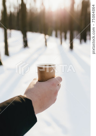 Guy holds paper cup of coffee in hand in the snowy forest. Blurred background 75864864