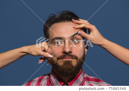 Female hands making different emotions of young bearded man on blue background Female hands making different emotions of young bearded man on blue background 75865348