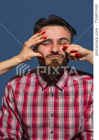 Portrait of young man with female hands punchning his face on blue background Portrait of young man with female hands punchning his face on blue background 75865349