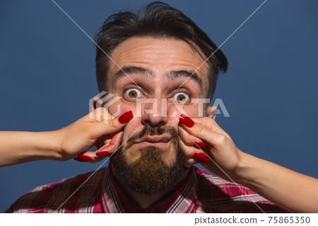 Portrait of young man with female hands punchning his face on blue background 75865350
