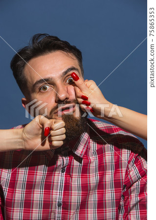 Portrait of young man with female hands punchning his face on blue background Portrait of young man with female hands punchning his face on blue background 75865353