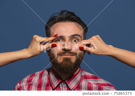 Female hands making different emotions of young bearded man on blue background Female hands making different emotions of young bearded man on blue background 75865373