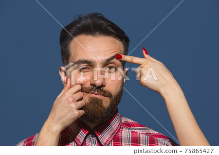 Female hands opening eye of young bearded handsome man on blue background 75865427