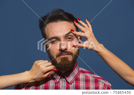 Female hands opening eye of young bearded handsome man on blue background Female hands opening eye of young bearded handsome man on blue background 75865430