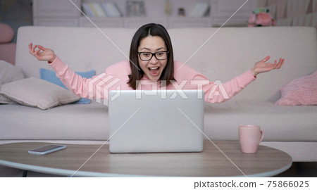 Excited overjoyed mixed race woman winner looks at laptop computer celebrates online success sits on sofa at home. Asian lady gets new distance job opportunity, reads good news, feels motivated. 75866025