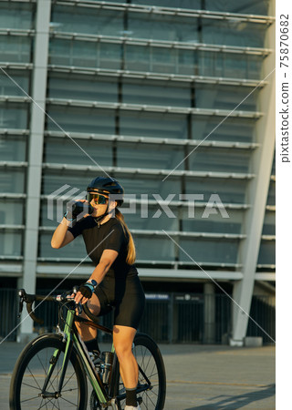 Professional female cyclist in black cycling garment and protective gear drinking water while riding bicycle in city, training outdoors on a warm day Professional female cyclist in black cycling garment and protective gear drinking water while riding bicycle in city, training outdoors on a warm day 75870682