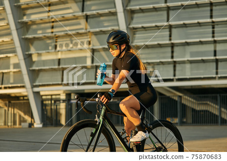 Professional female cyclist in black cycling garment and protective gear holding water bottle while riding bicycle in city, training outdoors at sunset Professional female cyclist in black cycling garment and protective gear holding water bottle while riding bicycle in city, training outdoors at sunset 75870683