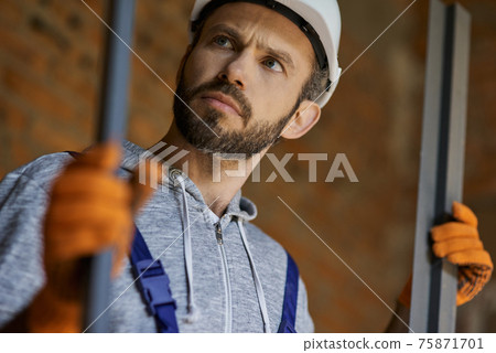 Close up shot of young male builder wearing hard hat looking focused, holding metal studs for drywall while working at construction site 75871701