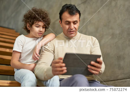 Middle aged hispanic father sitting on the stairs together with his son school boy, using digital tablet pc during remote studying at home 75872274
