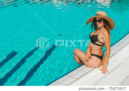 Female by pool. Happy young sexy girl in bikini swimsuit, sunglasses and straw hat in blue water. Luxury lifestyle travel. 75873917