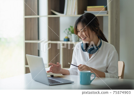 Portrait of a pretty young asian woman studying online with laptop computer while sitting at the table and notebook at home. 75874903