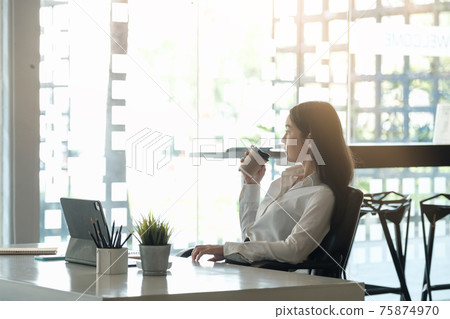 Portrait of beautiful woman or accountant sitting at desk in modern office with interior drinking hot beverage holding cup with coffee in hands looking outside enjoying freetime. 75874970