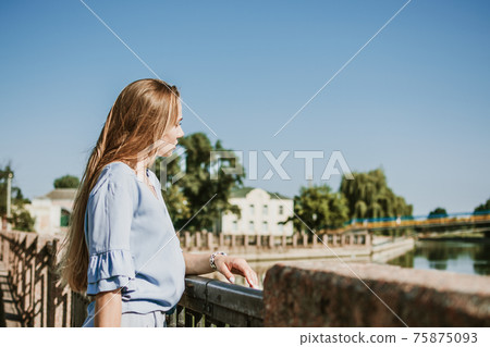 Lonely pensive Young blonde woman in blue dress standing near the city river in summer sun day. Alone deep in thought woman outdoors 75875093