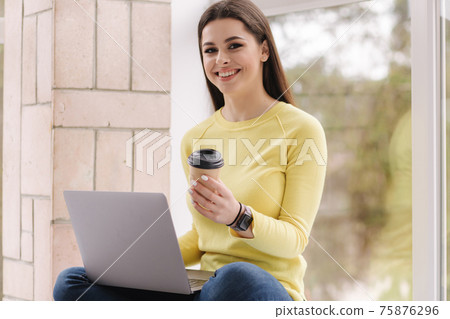 Portrait of attractive young woman using laptop in front of big window. Brunette woman in yellow sweatshirt working at home and drink coffee Portrait of attractive young woman using laptop in front of big window. Brunette woman in yellow sweatshirt working at home and drink coffee 75876296