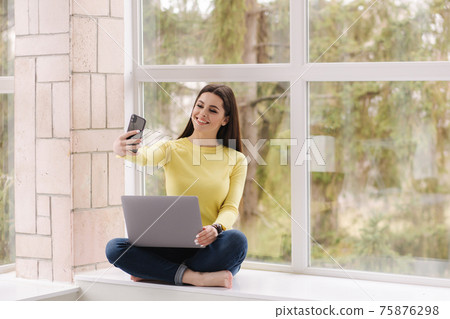 Young graphic designer working at home. Woman sitting on window sill with laptop and drink coffee. Self isolated female freelancer. Woman make selfie Young graphic designer working at home. Woman sitting on window sill with laptop and drink coffee. Self isolated female freelancer. Woman make selfie 75876298