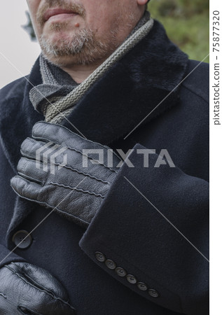 A cropped photo of a middle-aged man wearing a black coat and gloves. Adult male adjusting collar with hands. Soft focus. A cropped photo of a middle-aged man wearing a black coat and gloves. Adult male adjusting collar with hands. Soft focus. 75877320