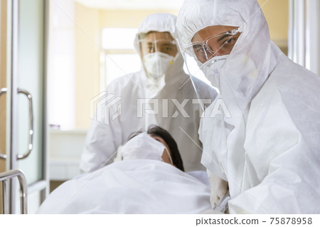 asian male nurse and doctor wearing ppe suit and face mask pushing stretcher gurney bed with seriously infected coronavirus or covid-19 patient towards quarantine room in hospital. 75878958