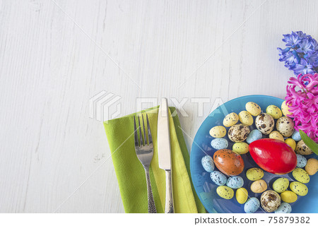 Festive place setting for easter holiday dinner. Blue plate with easter eggs, fork and knife on green napkin and blooming hyacinths on white wooden table top view. Space for text 75879382
