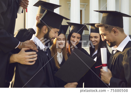 Group of happy students in mortar boards and bachelor mantle review the graduation album. Group of happy students in mortar boards and bachelor mantle review the graduation album. 75880508