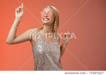 Delighted carefree attractive stylish millennial blond woman celebrating partying having fun wear sunglasses trendy silver dress dancing closed eyes broad smile waving hands up, red background 75885087