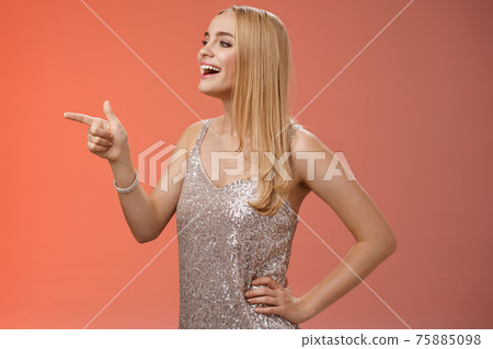 Stylish gorgeous young blond caucasian girlfriend in silver stylish evening dress turning profile left pointing curiously smiling broadly enjoying concert perfomance during party, red background 75885098