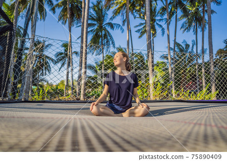 Young woman jumping on an outdoor trampoline, against the backdrop of palm trees Young woman jumping on an outdoor trampoline, against the backdrop of palm trees 75890409