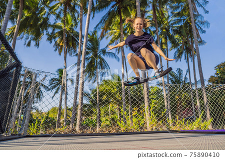 Young woman on a soft board for a trampoline jumping on an outdoor trampoline, against the backdrop of palm trees. The trampoline board is like a wakeboard, skateboard or snowboard trainer to hone 75890410
