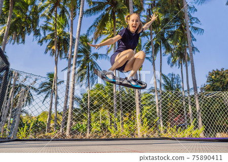 Young woman on a soft board for a trampoline jumping on an outdoor trampoline, against the backdrop of palm trees. The trampoline board is like a wakeboard, skateboard or snowboard trainer to hone Young woman on a soft board for a trampoline jumping on an outdoor trampoline, against the backdrop of palm trees. The trampoline board is like a wakeboard, skateboard or snowboard trainer to hone 75890411