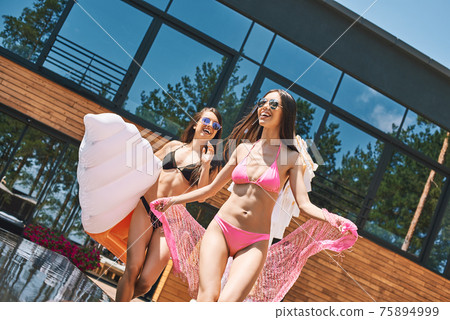 Enjoying summer vacation! Full length of two attractive young women in swimwear running near swimming pool together while one of them carrying ice cream shaped swimming mattress Enjoying summer vacation! Full length of two attractive young women in swimwear running near swimming pool together while one of them carrying ice cream shaped swimming mattress 75894999