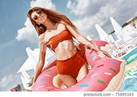 Beautiful one day, perfect the next. Sexy woman is wearing red bikini and holding inflatable swim ring in shape of donut Beautiful one day, perfect the next. Sexy woman is wearing red bikini and holding inflatable swim ring in shape of donut 75895165