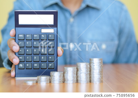 A businessman with a calculator and a stack of coins in foreground. Business, finance and growth A businessman with a calculator and a stack of coins in foreground. Business, finance and growth 75895656