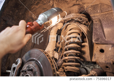 A car mechanic inspects shock absorber, springs and stabilizer bars with a flashlight. Car on a repair stand. Technical service station for car. 75896017
