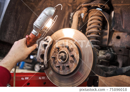 A car mechanic inspects brake discs and pads with a flashlight. Car on a repair stand. Technical service station for car. 75896018