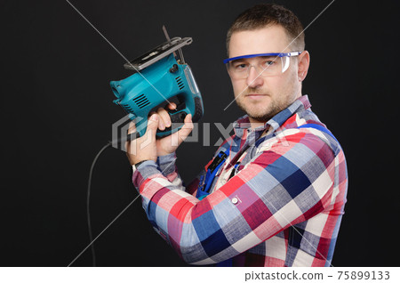 Caucasian man builder carpenter in a shirt and overalls with an electric tool in his hands. Studio portrait of friendly artisan businessman 75899133