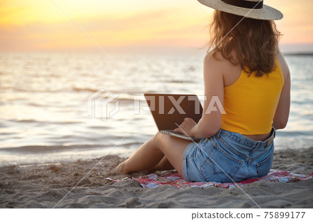 Young Caucasian Woman in shorts and a hat on the beach sitting on the sand browses social networks on the computer in the summer against the background of the sea. Remote work with a laptop 75899177
