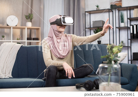 Joyful positive Arabian woman in hijab, using modern entertainment equipment, having fun on weekend at home, playing video games at living room, using VR glasses. Joyful positive Arabian woman in hijab, using modern entertainment equipment, having fun on weekend at home, playing video games at living room, using VR glasses. 75900961