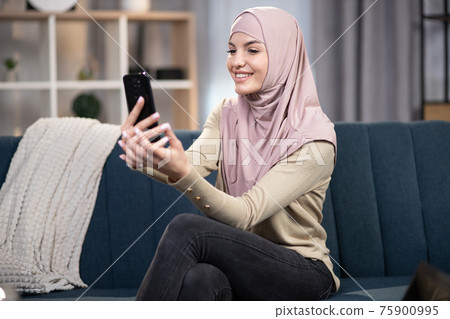 Video call, distance meeting, social networks concept. Pretty Arabian young girl in hijab, sitting on sofa at home, having video call using her smartphone, talking and having fun with friends 75900995