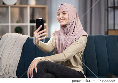Leisure at home, gadgets addiction. Pretty smiling Muslim girl wearing headscarf, sitting on comfortable sofa at home and using smartphone for making self photo 75900996