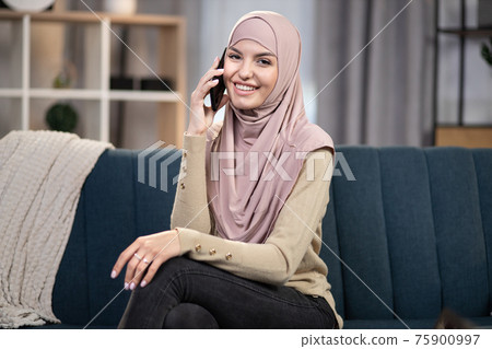Modern pleasant arabic girl in hijab, sitting on blue couch and talking phone with her friend, spending time at home, relaxing. People, gadgets and home leisure 75900997