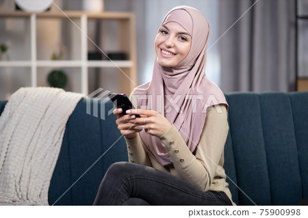 Young attractive smiling muslim woman, holding smartphone in hands, sitting on sofa couch, relaxing in living room at home, and looking at camera during leisure time 75900998