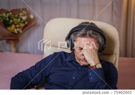 50-year-old man listens to music on headphones at home, sitting in a chair. 50-year-old man listens to music on headphones at home, sitting in a chair. 75902003